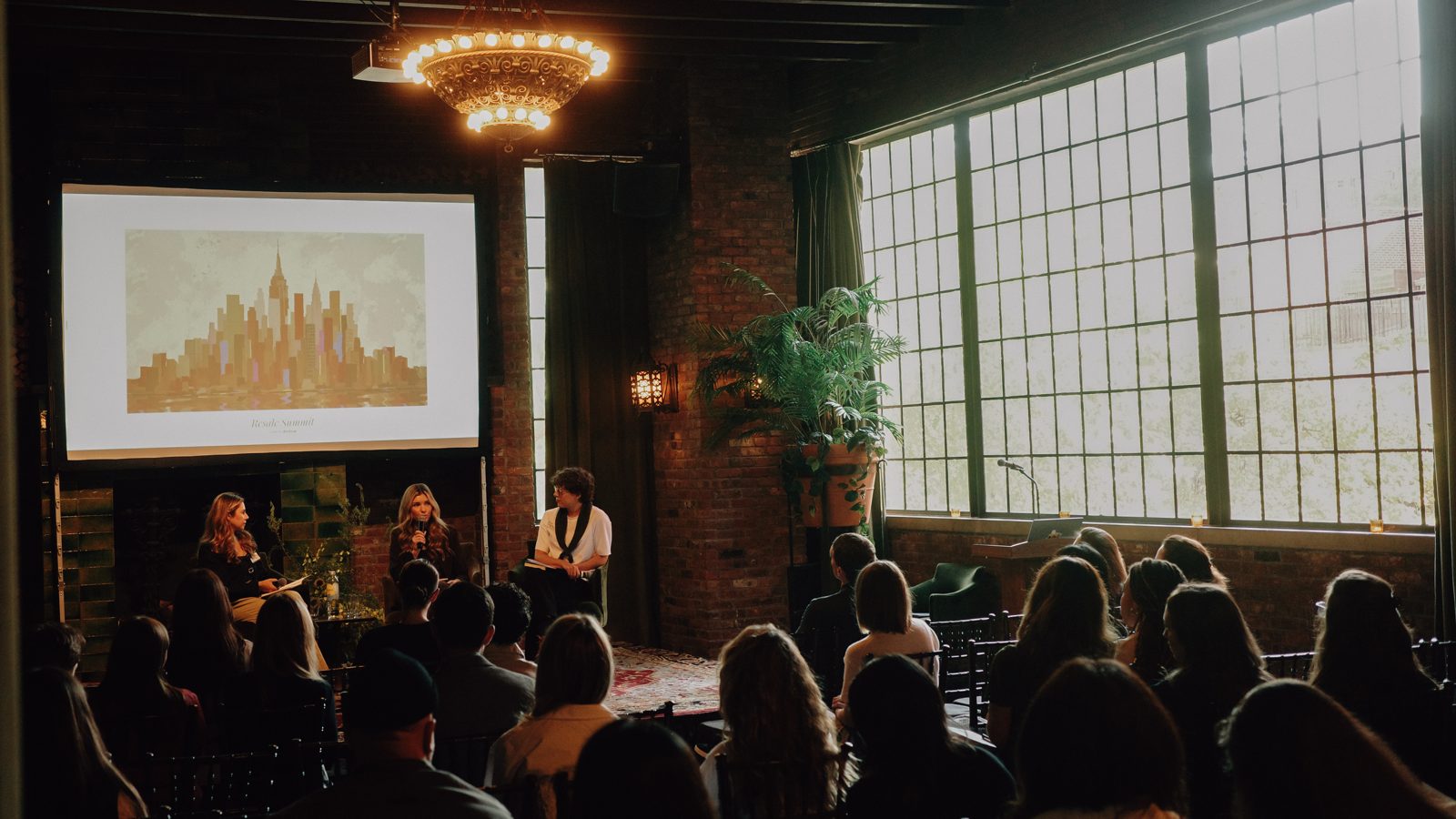 Three panelists seated on stage beneath a chandelier and a projected Resale Summit slide, with a full audience in the foreground and tall windows to the right